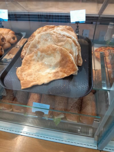The image shows a display case in a bakery or food establishment. In the foreground, a tray holds several golden-brown, flatbreads, some of which are folded. Behind the flatbreads, a partially visible pizza can be seen. Below this display, another shelf contains several sandwiches with sesame seed buns. The background shows more baked goods and a glimpse of a refrigerator unit. The overall environment suggests a commercial food service setting.