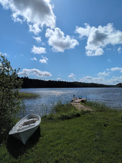 The image displays a bright, sunny lakeside scene, consistent with a natural landscape in Hartola, Finland. The sky occupies the upper portion, featuring a clear, light blue expanse adorned with scattered white cumulus clouds. The sun is high and to the right, casting intense glinting reflections across the rippled surface of the lake, particularly noticeable in the mid-left portion.
The lake itself is a broad body of water, bordered on the far side by a dense, dark green forest line that extends across the horizon. In the foreground, a vibrant green grassy bank slopes gently towards the water. A white wooden rowboat, containing two wooden oars, is beached on the grass on the left side of the frame. Towards the right, a wooden pier extends from the grassy shore into the lake, culminating in a small platform. A dark-colored chair or stool is positioned at the end of this pier. Patches of tall green reeds and other aquatic grasses grow along the immediate shoreline and around the base of the pier. A leafy green deciduous tree or large bush is partially visible on the far left foreground. Small yellow flowers are dispersed throughout the grassy area. No people or animals are present in the scene.