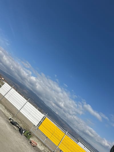 The image depicts an outdoor scene in the city of Razhdak, Bulgaria, captured with a significant counter-clockwise tilt. The upper two-thirds of the frame are dominated by a clear blue sky interspersed with white, stratocumulus clouds stretching horizontally across the view, particularly dense on the left. A distant landscape of rolling hills or mountains, appearing hazy and brownish-green, forms the horizon line. In the mid-ground, a long fence runs diagonally from the lower-left to the mid-right. This fence consists of metal mesh panels mounted on a concrete base with metal posts. The section of the fence on the left features white backing panels behind the mesh, while the section on the right features bright yellow backing panels. The foreground consists of a light brown, possibly gravel or dirt, ground surface. Resting on this ground, near the base of the white fence section, are several dark, irregularly shaped objects, possibly discarded plastic sheeting or fabric, alongside a rectangular green crate. Further to the right, near the yellow fence, a reddish-brown, circular basin-like object is visible on the ground. No individuals are present in the scene.