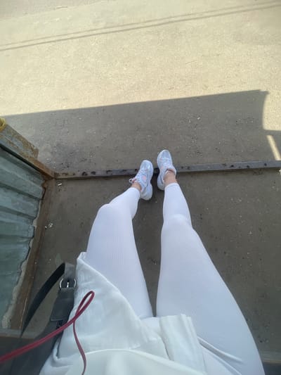 The image shows a person wearing white ribbed leggings and light grey and pink sneakers. They are standing on a concrete surface next to a rusty metal structure with holes, possibly a drain or part of a building. The person is also wearing a white garment, and a black bag with a red strap is visible. The specific location in the city of Molokovo, Russia, cannot be determined from the image.