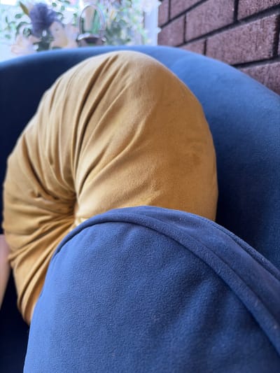 The image shows a close-up of a rounded, mustard-yellow cushion resting on a dark blue cushioned seat. The cushion has a gathered or ruched texture, creating deep folds that radiate from its center. The blue seat appears to be upholstered in a textured fabric. In the background, out of focus, a brick wall and some foliage are visible, suggesting an indoor or patio setting. The lighting is soft, and the overall impression is one of comfort and relaxation. The location is not specified in the image.