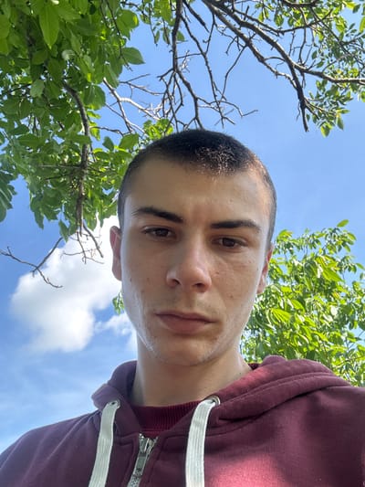 A low-angle shot depicts a person against a backdrop of trees, sky, and clouds. The person, likely male, has short, dark hair, brown eyes, and light skin, wearing a burgundy hooded jacket with white drawstrings and a visible zipper. The background includes tree branches with green leaves, and patches of blue sky with scattered white clouds. The photo's angle suggests the camera was positioned below the subject, looking upwards. The location in Oriahovo, Bulgaria, is not evident from the visual information provided.