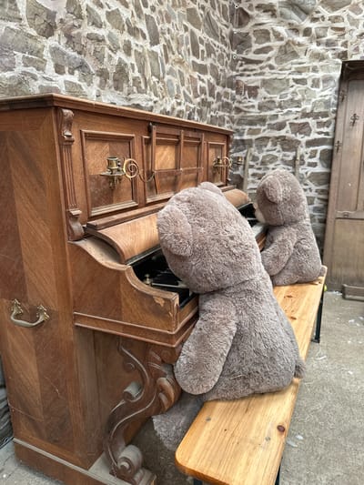 Two large teddy bears are seated on a wooden bench in front of an old, ornate wooden piano. The bear in the foreground is facing away from the camera, with its head angled towards the piano keys. The bear in the background is also facing the piano, with its head slightly turned. The piano is dark wood with intricate carvings and brass candle holders. The scene is set against a rustic stone wall, with a wooden door visible in the background. This image was taken in Gonneville-la-Mallet, France.