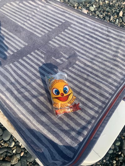 An overhead, high-angle shot captures a beach towel with alternating navy blue and white stripes, laid on a white surface next to a collection of smooth, dark grey pebbles. A transparent plastic bag containing a yellow, smiley-face-shaped candy wrapper rests on the towel. The wrapper is partially visible, revealing the design of a face with large blue eyes, a red mouth with a tongue sticking out, and text in Cyrillic script that appears to be "Сливочное" (Slivochnoye) and "Мороженое" (Morozhenoye), suggesting the candy is ice cream flavored. The shadow of an unseen object falls across the towel, indicating bright sunlight. The scene is set outdoors, likely at a beach in Gulripshi, Abkhazia.