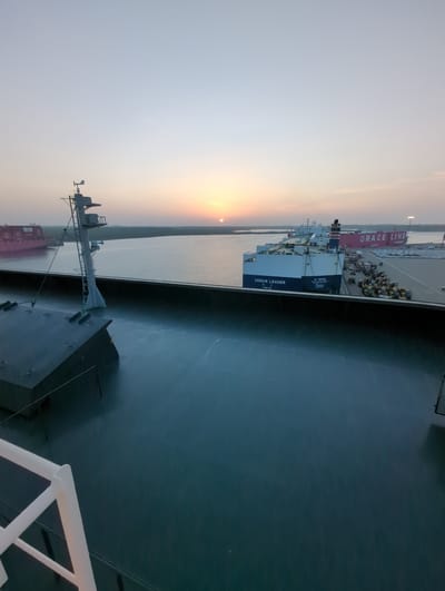 The image depicts a port scene at dusk in Hambantota, Sri Lanka. A large car carrier ship named "VENUS LEADER" is docked, laden with numerous vehicles, and another large pink ship labeled "GRACE LINE" is also visible. The sun is low on the horizon, casting a warm glow. The foreground shows the deck of a vessel, with a railing and a mast structure. The water is calm, reflecting the sky and the ships. The port appears to be a busy logistical hub.