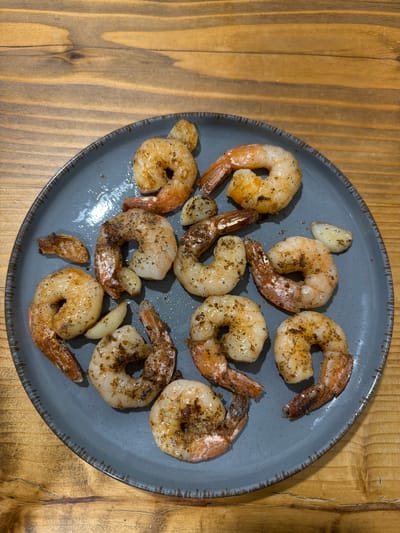 A plate of cooked shrimp and garlic slices sits on a wooden table. The shrimp are pinkish-orange and seasoned with pepper and herbs. Several pieces of garlic are scattered around the shrimp. The plate is dark gray with a textured rim. The wooden table has a visible grain pattern.