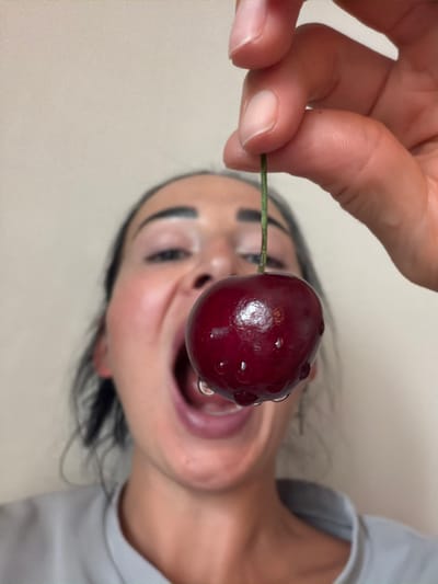 This is a close-up, rotated shot of a person holding a ripe cherry with a stem in their fingers. The cherry is dark red and has water droplets on its surface. The person's mouth is open, and they appear to be about to eat the cherry. The background is a plain, light-colored surface. The image's orientation suggests it might be a selfie or a photo taken by someone directly in front of the subject, with the camera rotated. The lighting is even, indicating it's likely an indoor setting with artificial light. There are no visible text elements in the image. Given the location context of Koltushi, Russia, this is likely a personal moment captured indoors. The emotion conveyed is one of anticipation or enjoyment of the fruit.