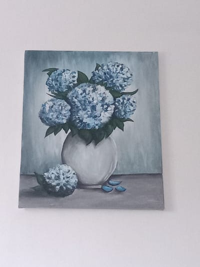 A painting of blue hydrangeas in a vase is displayed on a white wall. The painting is rectangular, oriented vertically. The hydrangeas are a vibrant blue, with some lighter and darker tones to create depth. They are arranged in a rounded bouquet within a light-colored vase, which sits on a grey surface. Below and to the left of the vase, a single fallen hydrangea bloom rests on the surface, along with a few small, blue petal-like shapes. The background of the painting is a muted, textured blue-grey. The painting is illuminated by ambient light, casting no discernible shadows. The context of the image being located in Kielce, Poland is not visually discernible from the painting itself.