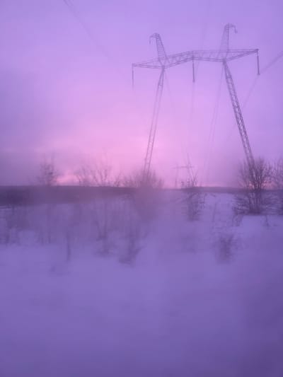 The scene depicts a winter landscape in Varenikovskaya, Russia, characterized by dense fog and a pervasive magenta-purple light. Two lattice-steel electricity transmission towers are visible in the upper portion of the frame; one large pylon occupies the mid-right, while a smaller pylon stands further in the distance to its left. Overhead transmission lines extend from these structures, disappearing into the thick fog. The foreground and midground consist of a snow-covered or frozen surface, possibly a body of water, which reflects the silhouettes of sparse, bare-branched trees and shrubs located along a low horizon line. Visibility is significantly reduced due to the heavy atmospheric conditions. No individuals, animals, or explicit actions are present within the frame.
