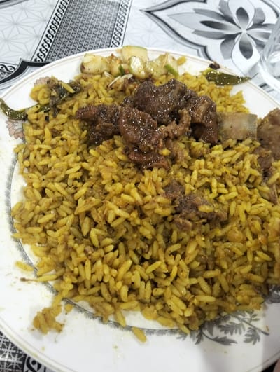 This image shows a plate of food, possibly beef biryani, set on a patterned tablecloth. The food consists of yellow rice and pieces of meat, likely beef, and is garnished with green chilies and a light salad. A glass is visible in the upper right corner of the image. The location, Naodoba, Bangladesh, is stated but not verifiable from the image alone. No people or interactions are visible.