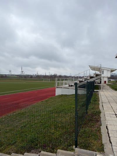 The image displays an outdoor sports complex in the city of Suvorovo, Bulgaria, under an overcast sky. The foreground features a dark green wire mesh fence separating a grassy slope from a cracked, light-colored paved pathway. To the left, a green artificial turf football field with white markings is visible, bordered by a red running track. Two white goalposts are present on the field in the mid-distance. To the right of the field and track, white concrete spectator stands with railings and safety netting rise, featuring an elevated white booth with a pointed roof and antenna at its upper right end. In the background, a line of bare trees is visible, with multiple large metal lattice power line towers extending across the horizon. No individuals are present within the scene.