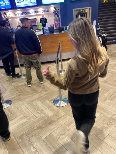 People are standing in line, presumably indoors on a herringbone-patterned floor. A woman with blonde hair wearing a tan jacket and black pants gives a thumbs-up. A man in a black jacket and khaki pants is standing in front of her. Another person in dark clothing is at the front of the line. In the background, there's a counter and promotional material for Topgolf Miami Doral. Golf clubs are also visible.