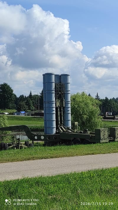 In the city of Лашанскі сельскі Савет, Belarus, a military missile system is displayed outdoors on a sunny day with scattered clouds. The missile system consists of four vertical cylindrical missiles mounted on a trailer. The missiles are light blue and are stacked in two pairs. The trailer is olive green and has multiple wheels. In the background, there are trees, a body of water, and a small blue building. A paved path runs in the foreground. The image is watermarked with "REDMI NOTE 9 PRO AI QUAD CAMERA" and "2026/7/15 10:29".
