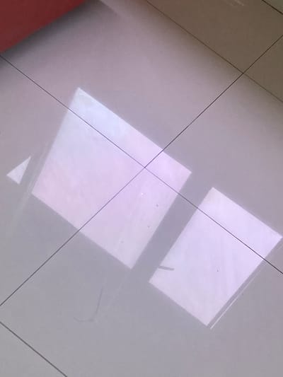 The image shows a tiled floor with two rectangular reflections of light. The tiles are square and arranged in a grid pattern, with thin dark grout lines. The reflections are bright and pinkish-white, suggesting they are from a light source such as a window or a lamp. There is also a small triangular reflection in the upper left quadrant. The upper portion of the image features a blurry red object, possibly furniture or a wall. The floor appears to be clean. There are no people or animals visible in the image. The image is taken from a high-angle perspective, looking down at the floor. The location is stated to be in the city of Proyonangg, Indonesia.