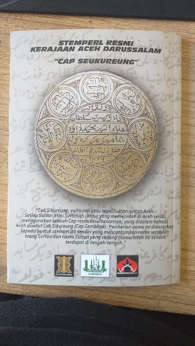 The image displays the cover of a document or book, centrally positioned on a light-colored wooden surface. A dark, undefined object, possibly a keyboard or mat, partially obscures the upper left portion of the wooden surface. The cover is predominantly off-white with faint background Arabic script. Bold black text at the top reads "STEMPERL RESMI KERAJAAN ACEH DARUSSALAM" followed by ""CAP SEUKUREUNG"". A large, ornate circular emblem, appearing gold or metallic, dominates the center, featuring intricate Arabic calligraphy within a central circle and eight surrounding circular segments. Below the emblem, a paragraph in black Indonesian text describes "Cap Sikureung" as the official seal of Acehnese Sultans, noting it incorporates the names of nine Sultans, with the reigning Sultan's name centrally located. Three rectangular logos are aligned horizontally at the bottom: a golden, heraldic design on the left; a green and white logo depicting stylized minarets and "ACEH DARUSSALAM" in the middle; and a red and black logo showing crossed traditional swords on the right. The scene is situated in an indoor environment within Batang Kuis, Indonesia.