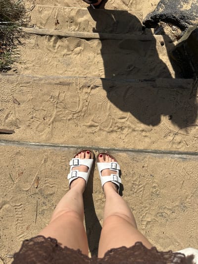 From a high angle, a person stands on sandy steps, wearing white Birkenstock sandals. Their toes are painted a deep red. The steps are lined with what appears to be metal or concrete edges. The lower left corner of the image shows grass and a small portion of another shoe. A dark, indistinguishable shadow falls across the steps, and a portion of what seems to be a brown, lace-like garment is visible on the right. The sandy surface of the steps is marked with footprints.