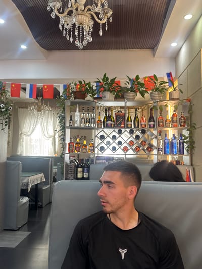 The image depicts an interior scene within a restaurant in Suifenhe, China. A young man with short, dark hair and a light beard is seated on a grey booth, looking to his right. He is wearing a black t-shirt with a white symbol on the left chest. Another person, partially visible and with dark hair, is seated behind him. The restaurant features a decorative chandelier hanging from a dark, ribbed ceiling. Along the back wall, there are shelves stocked with various bottles, including what appear to be spirits and wines. A wine rack with multiple compartments is also visible. In the background, curtains are drawn across a window, and there are flag decorations, including Chinese flags, hanging from the ceiling. Tables with tablecloths are set up in the dining area. The overall environment suggests a casual dining establishment.