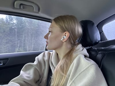 The image shows a young woman in the passenger seat of a car, looking out the window. She has blonde hair and is wearing a light-colored hooded sweatshirt. She has white earbuds in her ears. The car appears to be in motion, with trees visible through the window. The interior of the car is visible, including a headrest and a grab handle. The scene is likely in Didino, Russia, given the context.
