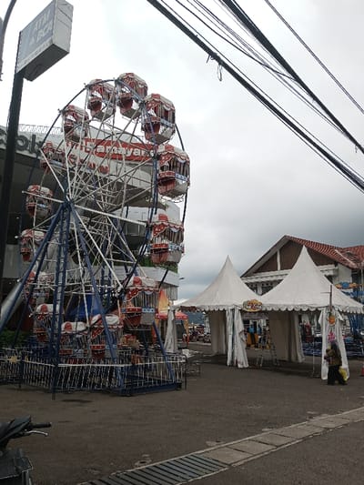 A Ferris wheel stands tall, its red and white cars contrasting against the overcast sky. Power lines stretch overhead. The wheel is near a building and set up on a street next to tents with peaked roofs. A metal fence surrounds the base of the Ferris wheel. The street is made of asphalt, and a drain is visible on the edge of the street. Part of a motorcycle is visible in the lower left corner.
