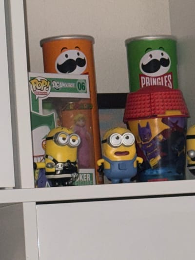 A shelf holds two Pringles cans, green and orange, alongside a Batman-themed container. Two Minion figures are also present: one in overalls and another chained, possibly a Joker variant, standing in front of a green DC Heroes Funko Pop box. The setup is cluttered, and the objects appear to be toys or collectibles.