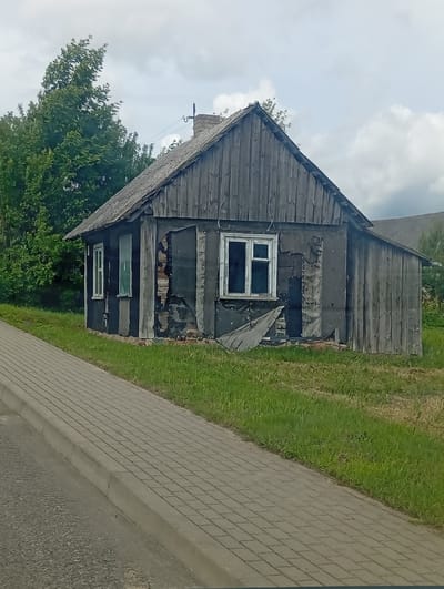 The image depicts a dilapidated wooden house located in Šaukėnai, Lithuania. The house is situated in a grassy area next to a paved sidewalk and a road. The wooden siding of the house is weathered and aged, with some sections showing damage and peeling. There are two windows visible on the front of the house, one with white trim and glass, and another with a green tinted pane. A chimney is visible on the roof. Lush green trees are present behind the house. The sky is overcast with clouds. The scene suggests a rural or semi-rural setting with a focus on the decaying structure.