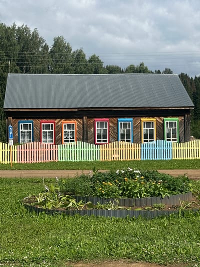The image shows a long, wooden house with a corrugated metal roof. The house has six windows, each with a colorful frame: blue, red, orange, pink, blue, and green. A rainbow-colored picket fence runs in front of the house. To the left of the house, there is a blue sign with a white "P" symbol and a wheelchair accessibility symbol. In the foreground, there is a circular flowerbed with various plants and flowers. The scene appears to be in a rural setting, possibly Zapadnaya, Russia, with trees in the background and a dirt road in front of the flowerbed. The sky is overcast with some clouds. No people are visible in the image.