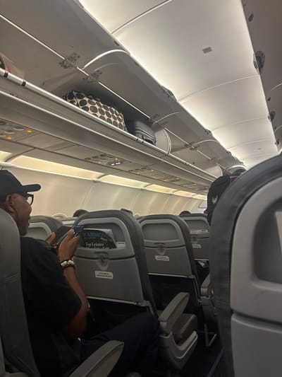 An eye-level indoor shot from inside an airplane cabin depicts a male passenger seated and using a smartphone. He is wearing glasses, a black hat, a black shirt, and a bracelet. Overhead compartments are visible above the seats, and some contain luggage. One magazine rack contains reading material with "Top Leisure" visible on its cover. The cabin is generally brightly lit with overhead lighting. There is no evidence in the image to support that this image was taken in Omoecheigbo, Nigeria.