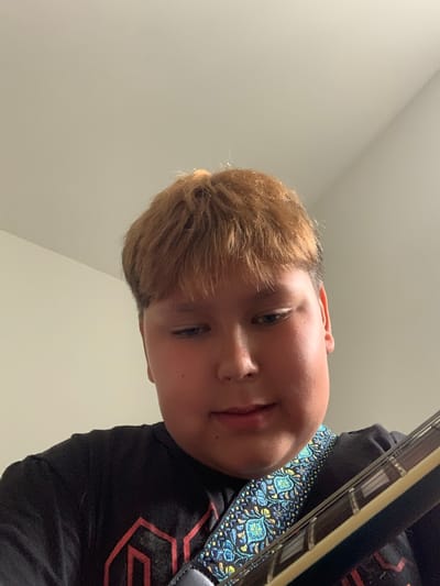 A young male with reddish-brown hair and light blue eyes is shown from the chest up, looking down. He is wearing a black t-shirt with a red geometric design. A guitar strap with a blue, yellow, and black pattern is visible across his chest, and the neck of a guitar is in the foreground. The background is a plain, light-colored wall. There is no visible indication of the location being in Vejprty, Czechia.