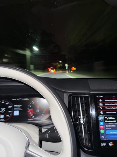 The image depicts a first-person perspective from the driver's seat of a vehicle at night, likely in motion, on Edgebrook Road in Lawrence Township, New Jersey, United States.
The foreground shows the interior of a modern vehicle. A cream-colored steering wheel partially obstructs the view of the digital instrument cluster, which displays a speed of "35" units, a fuel gauge near full, and other vehicle information. To the right of the steering column, a wiper stalk is visible, set to "INT" (intermittent). To the right of the dashboard, a vertical air vent with a silver adjustment lever is present, beside a large infotainment touchscreen. The screen displays an active Apple CarPlay interface with "Guidance via Apple Car...", "Phone via Apple CarPlay", "Slack's Phone", and navigation showing "Edgebrook Rd" with "Construction" ahead at "2.2km". The time displayed is "10:05".
Through the windshield, the dark road ahead is visible, illuminated by streetlights and the vehicle's headlights. The windshield has streaks, suggesting it is wet or has recently been wiped. A vehicle's red taillights are visible directly ahead on the road. The road appears to be asphalt with a yellow dividing line. Streetlights are visible on both sides of the road, and the surrounding environment consists of dark foliage, likely trees or bushes, in a suburban or residential setting.