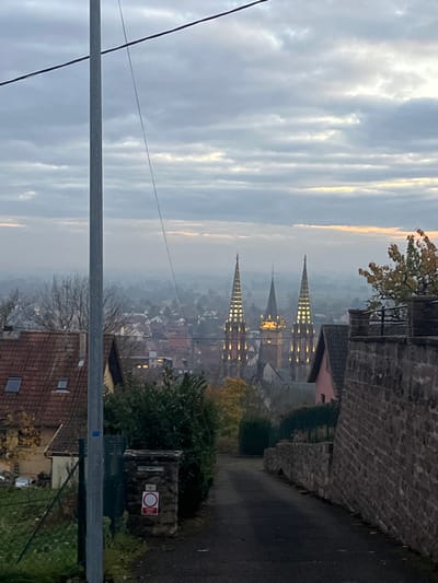 The image displays a panoramic view of the city of Obernai, France, under an overcast sky during what appears to be either dawn or dusk. The sky is largely grey with horizontal cloud formations, exhibiting faint orange and yellow light near the horizon.

In the foreground, a dark asphalt path slopes downwards, bordered on the left by a tall grey utility pole from which dark wires extend across the upper frame. Adjacent to the pole, a green metal fence separates the path from a residential property featuring a house with a red tiled roof and two skylights. Further along the left side of the path, a stone pillar bears the number "3" and a circular red prohibition sign with a white horizontal bar. Dense green hedges line this section of the path.

On the right side of the foreground, a robust stone wall constructed from irregularly shaped, reddish-brown blocks runs parallel to the path, also sloping downwards. A tree with some yellowing autumn leaves is visible above this wall.

The midground reveals additional houses with varied rooflines nestled amongst trees and hedges. The path continues into the distance, descending towards the city center.

Dominating the background are the illuminated spires of a prominent church, specifically the Église Saints-Pierre-et-Paul in Obernai. Two tall, slender spires on the left and right feature multiple bands of golden light, while a central, darker spire is positioned slightly behind them. Beyond these illuminated spires, a dense urban landscape of residential buildings with red roofs extends into the hazy distance, characteristic of the city of Obernai. A pervasive atmospheric haze or light fog obscures distant details. No individuals are visible in the scene.