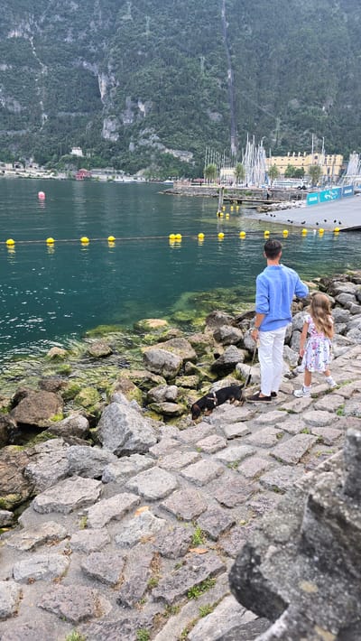A man, a young girl, and a dog are walking along a rocky shoreline on a cloudy day. The man is wearing a blue long-sleeved shirt and white pants, and he is holding the dog's leash. The girl is wearing a floral dress and white shoes. The dog is small and black with some brown markings. The water is a deep green, and there are yellow buoys floating in it. In the background, there are steep, tree-covered hills and buildings that appear to be part of a town. A marina with sailboats is also visible. The scene suggests a leisurely walk by the lake. The location is Riva del Garda, Italy. No visible text in this image.