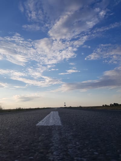 The image depicts an asphalt road leading into the distance under a vast, partly cloudy blue sky. A white line is painted on the road, marking the lane. In the far distance, a signpost stands on the left side of the road, and a line of trees is visible on the right horizon. The foreground of the road is dark and textured, suggesting it is made of gravelly asphalt. The sky is a vibrant blue, with scattered white clouds, some wispy and others more substantial. The overall impression is one of an open, rural landscape. The location is identified as Dölen Rural District, Kazakhstan.