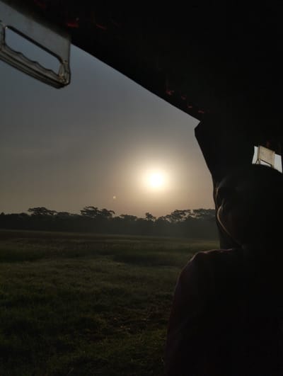 The image depicts a landscape at sunrise in Southkhali, Bangladesh, as viewed from inside a vehicle. The sun, a bright orb in the sky, casts a hazy glow over a field of grass in the foreground and silhouettes trees on the horizon. The interior of the vehicle is dark, obscuring details, but a portion of a person's head and shoulder is visible in silhouette on the right side of the frame. A metallic handle is visible above, against the muted sky. The overall scene suggests a tranquil, early morning moment captured from within a moving vehicle.
