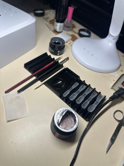 The image displays a collection of nail art supplies on a light-colored, textured surface. In the upper left is a white UV nail lamp with several buttons. To its right, there are two small black containers with lids, one open and revealing a gel-like substance, and the other closed. A pink-capped bottle of nail polish remover is also present.

Central to the image is a black organizer holding several nail art brushes with red and black handles, and a wooden cuticle pusher. Next to this, another black organizer contains a set of five clear plastic nail tips.

In the lower right, a small pair of silver scissors with rounded tips is visible, alongside a black electrical cord. A small white paper pad is situated to the left of the nail brushes. The lighting suggests an indoor setting, possibly a home or salon workspace. The background is out of focus, with hints of dark surfaces and patterned elements. No people or animals are visible in the image. The scene is static, with no ongoing actions. The objects appear to be neatly arranged for use. The specific location of Yubileynoye, Russia, cannot be determined from the visual information provided in the image.