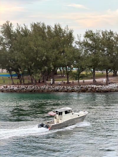 A medium shot captures a boat with "UAP RESPONDER" written on its side, moving through green-tinted water, leaving a white wake. The boat, with twin outboard motors, displays an American flag. In the background, a rocky shore separates the water from a park area with picnic tables, trees, and a few people. The sky above is a pale gradient of pink and blue, with a faint distant airplane. The scene appears to be a coastal area in Bal Harbour Village, Florida, United States of America.