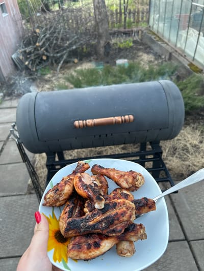A plate of grilled chicken sits next to a vertical smoker grill on a gray concrete patio. A hand with red nail polish holds the plate, which has a floral design. The grill is black with a wooden handle. In the background, there's vegetation, possibly a garden or lawn. The scene appears to be outdoors. There is no evidence of the location being Bashur, Russia.