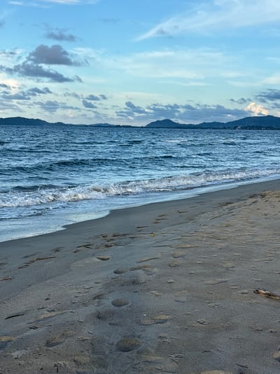 The image depicts a coastal scene in Tela, Honduras, featuring a sandy beach, ocean, distant land, and sky. The foreground comprises a grayish-tan sand beach occupying the lower two-thirds of the frame. Numerous shallow human footprints are visible across the sand. A small, round yellow object is located on the sand in the mid-left, and a piece of driftwood or log lies on the far right edge of the sand. The Caribbean Sea occupies the midground, with moderate waves breaking into white foam along the shoreline. The water exhibits varying shades of blue and green. In the distant background, a series of dark, undulating hills or mountains covered in vegetation are visible across the water. The sky above is a light blue with scattered white and gray cumulus clouds. No individuals are present in the scene.