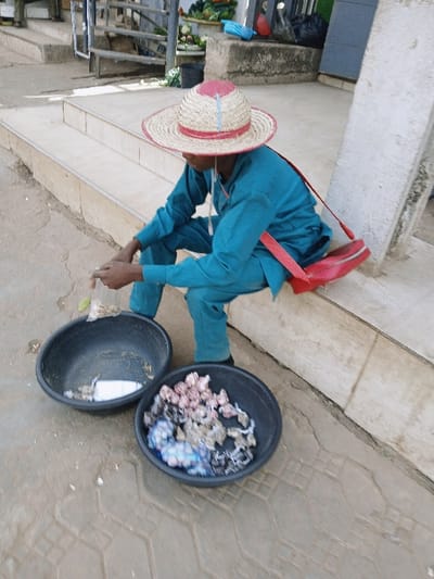 An individual is depicted seated on a concrete step in an outdoor commercial area of Mumbwa, Zambia. The person wears a wide-brimmed straw hat with red and light blue trim, a teal-colored long-sleeved shirt, and matching trousers. A red bag with a shoulder strap is slung over the right shoulder. The individual is looking downwards, holding a small clear plastic bag containing a white substance. On the ground directly in front of the person are two black plastic basins. The basin closer to the individual contains a small quantity of a white substance and some indistinct items. The second basin, positioned further away, holds numerous small, irregularly shaped items, some wrapped in colorful materials like pink, blue, and white. The ground beneath the basins is paved with interlocking concrete bricks. In the background, additional concrete steps ascend to a higher level, revealing a market-like setting with produce, a metal railing, and other objects on a concrete ledge.