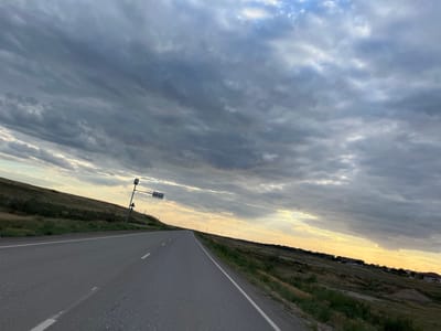 The image depicts a landscape scene, seemingly at dusk or dawn due to the soft, golden light illuminating the sky. A paved road with white lane markings stretches into the distance, curving slightly to the right. The road is bordered by a grassy embankment on the left, which appears somewhat wild and untamed, with some scattered structures visible in the far distance. On the right side of the road, the embankment is also grassy but appears more cultivated or maintained. The sky is filled with a dramatic display of clouds, ranging from dark, heavy masses to lighter, wispy formations, catching the golden light. A traffic signal is visible on a pole near the road, suggesting this is a public thoroughfare. The overall mood is serene and vast, with a sense of open space and natural beauty. The orientation of the image is rotated 90 degrees clockwise. The location is identified as Ilyinka, Russia.