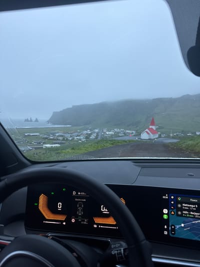 The image, taken from inside a vehicle, shows a view through the windshield of the town of Vik, Iceland. It is a rainy day. Visible outside the vehicle is a landscape featuring a village, green hills, and a coastline with sea stacks. A church with a red roof stands prominently within the village. The vehicle's dashboard is visible, displaying a digital instrument cluster indicating the car is in electric mode. The navigation system shows directions towards Klettsvegur 5, 1.7km away, and Höfn - Berjaya, 272km away. The time is 22:25, and the outside temperature is 8°C. The vehicle has travelled 6139km.