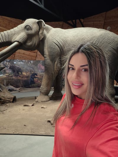 A woman is taking a selfie in front of a diorama of prehistoric elephants. The diorama depicts a scene with two mammoths and a bear in a wooded environment, with a grassy ground. The woman is wearing a red sweatshirt and has long, blonde-brown hair. The background consists of a wooden wall and ceiling, with a dark ceiling structure. The diorama is located in the Dorkovo, Bulgaria paleontological museum.