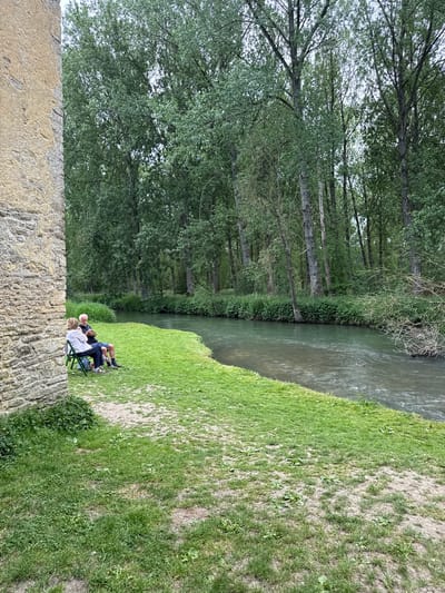 A sideways shot shows a riparian environment in West Oxfordshire, United Kingdom, with a tree-lined river on the right. A stone wall occupies the lower left of the frame. On the right, a grassy bank slopes down to the river's edge. Two individuals are seated on folding chairs at the edge of the bank, seemingly enjoying the view. One wears a light-colored shirt, and the other a dark shirt. The image captures a tranquil outdoor scene.