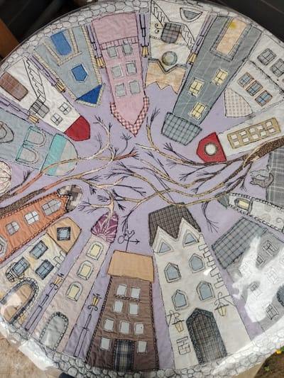 This is a close-up, circular, mixed-media textile artwork, likely a quilted table runner or placemat, depicting a stylized, whimsical cityscape. The dominant subjects are various buildings, rendered with fabric patches and intricate stitching. These buildings are arranged radially, radiating outwards from a central element of bare tree branches.

The buildings themselves are diverse in design and color, featuring patchwork walls in patterns like stripes, plaids, and checks, with fabric "roofs" and fabric "windows" outlined in black stitching. Some buildings have arched doorways and decorative architectural details. Scattered amongst the buildings are street lamps, also created from fabric and stitching, and a few small, stylized objects like a bicycle and a cat. The background fabric is a soft lavender. The overall impression is one of a charming, slightly surreal, European town.

The artwork is likely intended to evoke a sense of community and urban life, presented in a handcrafted, artistic manner. The time of day and weather are not explicitly indicated, but the gentle lighting of the street lamps might suggest an evening or early morning scene. The location context of Saint Petersburg, Russia, hints at the architectural style, which can be reminiscent of European cities. There is no visible text in the artwork itself. The artwork is framed by a clear plastic protective layer, suggesting it is a functional item.