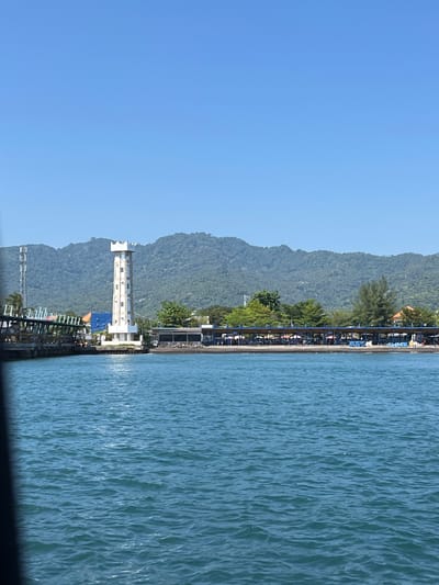 The image depicts a view of Pemenang Timur, Indonesia, from the water. The scene includes a clear blue sky, a forested coastline, a pier, and the ocean. A tall, white building is visible along the coast. The pier extends into the water, with structures and objects visible on it. The water appears calm with minor ripples.