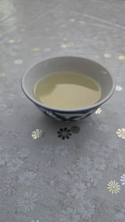 A close-up, slightly angled shot shows a white ceramic bowl filled with pale yellow liquid, likely tea or a similar beverage. The bowl has a blue, decorative pattern around its rim, featuring what appear to be birds or floral motifs. It sits on a tablecloth with a repeating floral pattern in white and gold on a textured gray background. The image is static and does not depict any specific location within Samarkand, Uzbekistan, nor does it show any people or actions.