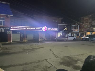 The image shows a street scene at night in Khasavyurt, Russia. A brightly lit sign in Russian reads "ЦЕНТР КРАСОТЫ & ЖЕНСКАЯ КОФЕЙНЯ" which translates to "CENTER OF BEAUTY & WOMEN'S COFFEE SHOP." Next to it is a logo with a stylized woman and the word "ЯСМИНА" (Jasmine). The building features a second story with windows and an external staircase. Several cars are parked on the street and in front of the establishments. The street surface appears wet and somewhat uneven, suggesting recent rain or damp conditions. The overall atmosphere is commercial and urban, illuminated by artificial lighting.