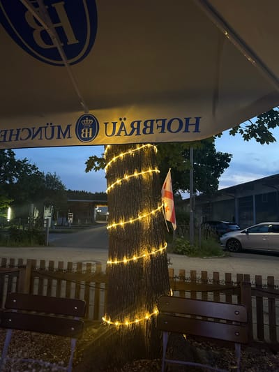 An outdoor seating area is depicted at dusk, likely at a beer garden or restaurant, with a large umbrella in the foreground displaying the Hofbräu München logo. A tree trunk wrapped in warm-toned string lights is a central element. A Georgian flag is attached to the tree. In the background, there are wooden benches, a fence, some vehicles parked along a road, and buildings that appear to be industrial or commercial. The sky is a twilight blue with some clouds. The overall atmosphere suggests a relaxed evening setting. The Hofbräu logo on the umbrella strongly suggests this location is connected to the Hofbräuhaus brewery, which has locations in various places, including Bad Endorf, Germany.