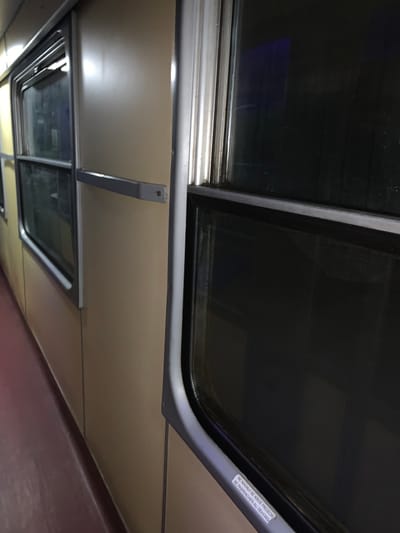 A view looking up at the ceiling of a train car with several rectangular windows. The window frames are gray, and the glass is dark. A sign is attached to one of the frames that reads "NE NAGINSE SE KROZ PROZOR, NE PASPENCHER AU DEHORS" in multiple languages. The ceiling panels are light brown, and metal supports extend down to hold up the window frames. One window is in the foreground, and another is visible further away.