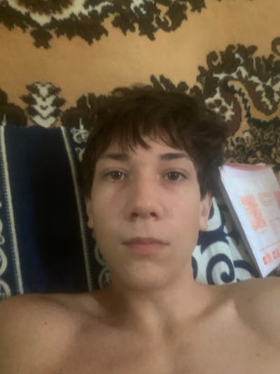 A close-up, high-angle selfie of a young male appears to be taken in Khavast, Uzbekistan. The subject has short, dark brown hair and is looking directly at the camera. He is shirtless, revealing his shoulders and upper chest. He is lying down, with a patterned blanket or rug visible behind him. The blanket features a brown and tan abstract design. To the right of the subject, a white envelope with red writing is visible. The image appears to be taken indoors, likely in a bedroom, given the presence of bedding and the intimate nature of the selfie.