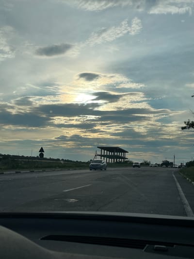 The image captures a scene along a road, likely in Vadul lui Voda, Moldova, with a dramatic sky overhead. The sun, partially obscured by clouds, casts a warm, golden light, suggesting it's either sunrise or sunset. The sky is filled with a variety of clouds, from wispy, elongated formations to darker, more substantial masses, creating a dynamic and textured ceiling.
The road itself appears to be a paved surface with clear lane markings, indicating it's a route for vehicular traffic. Several cars are visible, driving in the same direction, suggesting a journey or travel. A modern, open-sided shelter structure stands beside the road, its dark frame contrasting with the bright sky. In the foreground, the edge of a vehicle's interior is visible, with a dark, textured surface, implying the photo was taken from inside a car.
The surrounding landscape is green and appears to be covered in vegetation, with trees visible in the distance. Traffic signs are also present along the roadside, providing navigational cues. The overall mood is one of transit and observation, with the striking sky being a prominent feature. There is no visible text in the image.